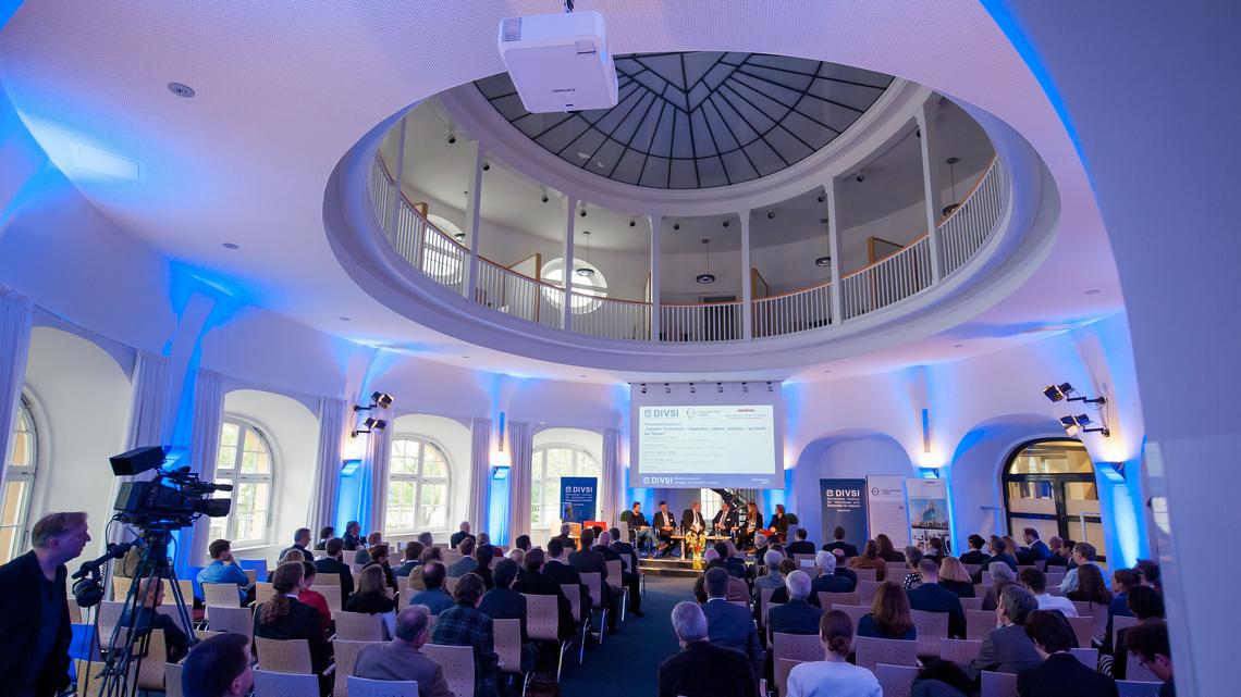 A view from behind: looking out on the Moot Court from the rear of the room, participants are seated in rows while a panel in engaged in discussion on the stage at the front of the room; blue LED spots have been used to illuminate the walls