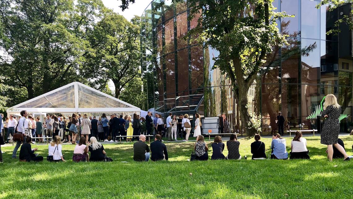 A view of the campus courtyard while an event is being held: in the foreground, students and guests are visible from the back - they are seated along one of the terrace edges while a crowd is gathered in- and outside of a tent that has been connected to the auditorium to provide additional space for catering