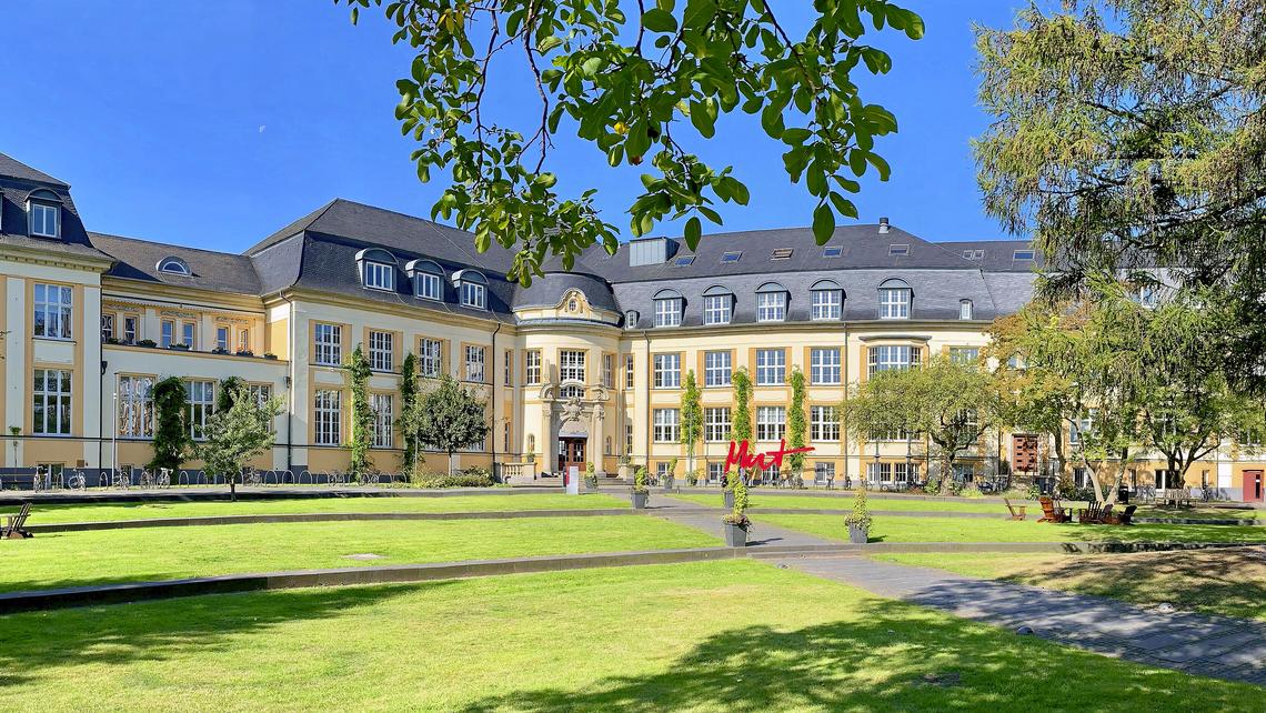 A photo from the Auditorium: looking out over the campus courtyard, the historic central building is framed by leafy branches from trees on either side, a strikingly blue sky over the curved roofline and bright green grass along the terraced walkway.