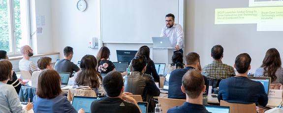 Students seated in a classroom take in a presentation; a slide is projected on the wall in the background while the lecturer delivers points from the front of the room