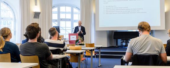 Seated in the Moot Court, participants take in a lecture: the presenter is gesticulating from a podium while a slide is projected on the screen behind him